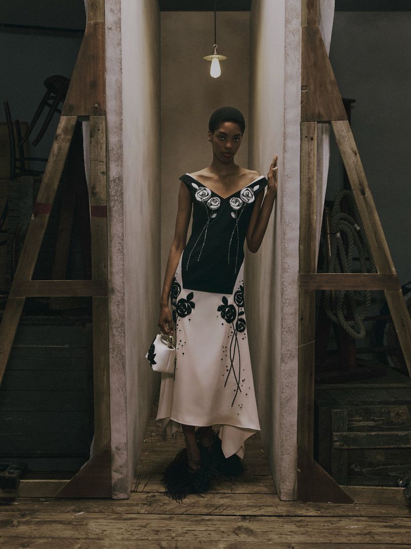 Model standing between wooden frames wearing a black and white floral dress holding a small white handbag.