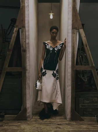 Model standing between wooden frames wearing a black and white floral dress holding a small white handbag.