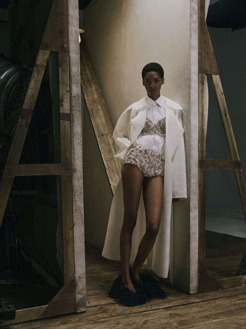 Model Aluel Keror standing between studio flats wearing an open white trench coat, collared shirt, and embellished high-waisted shorts.