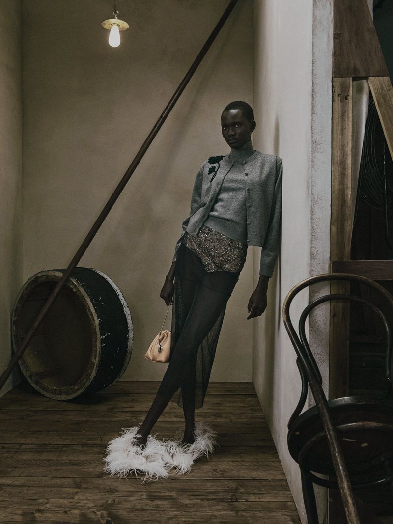 Model leaning against a wall in a grey knit cardigan and sheer skirt with feathered shoes for Erdem Pre-Fall 2027.