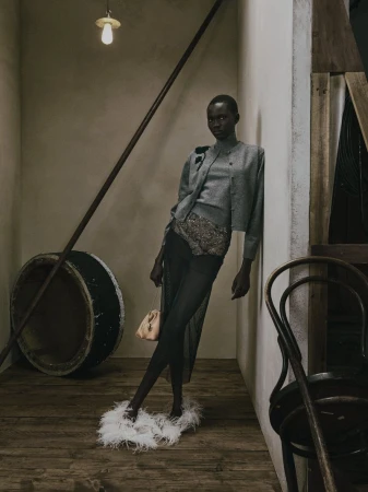 Model leaning against a wall in a grey knit cardigan and sheer skirt with feathered shoes for Erdem Pre-Fall 2027.