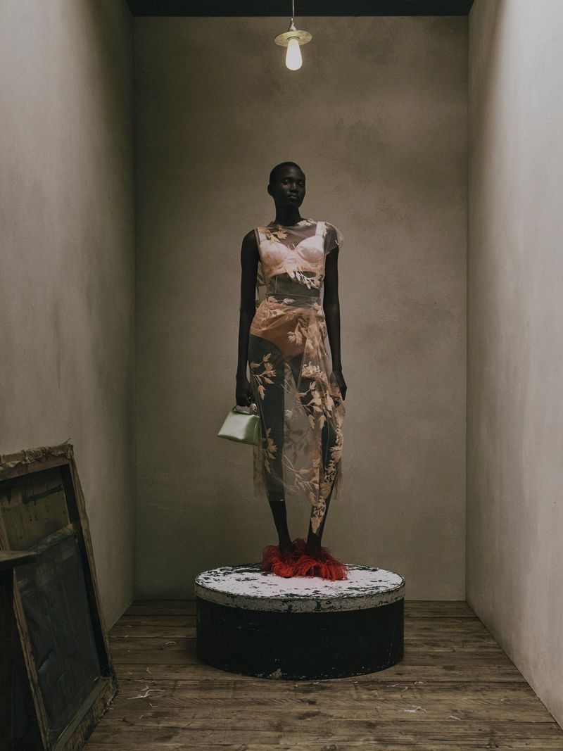 Model Aluel Keror stands on a circular plinth wearing a sheer floral embroidered dress and red feathered shoes under a hanging bulb.