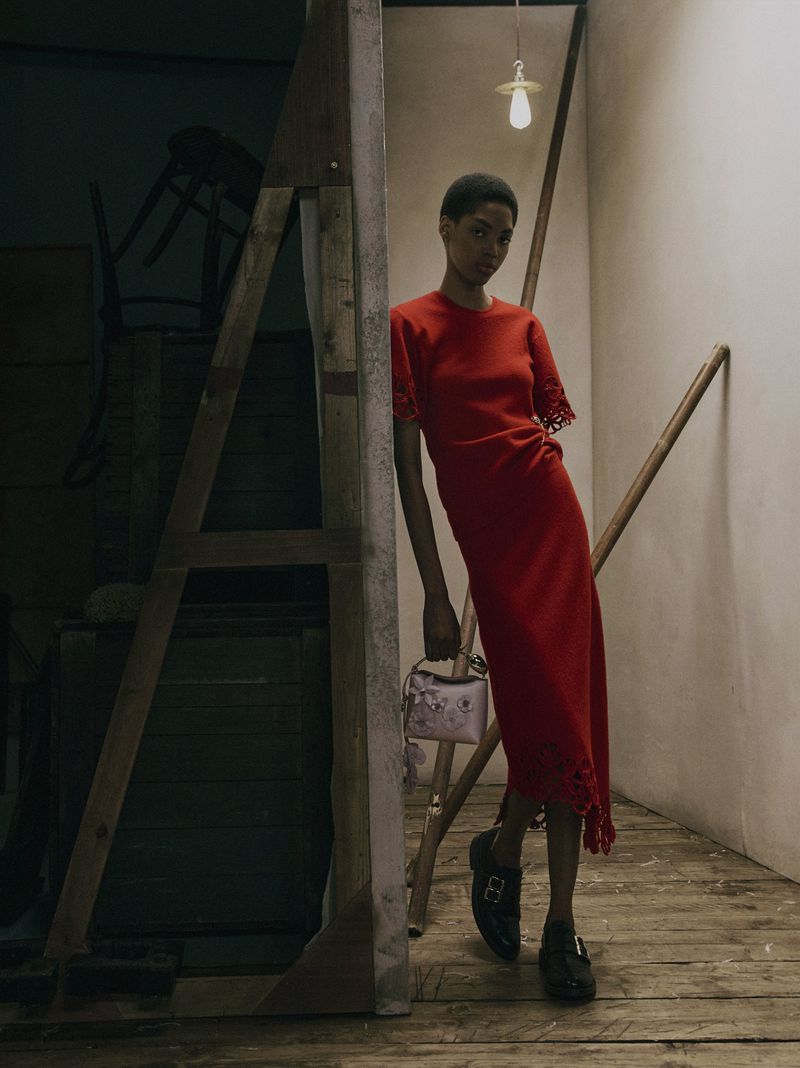 Model posing in a red short-sleeve midi dress holding a small pink handbag within a studio set of wooden beams.