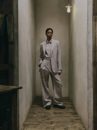 Model Aluel Keror in a tailored light grey pantsuit standing in a narrow plaster alcove with rustic wood floors.