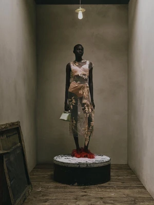 Model Aluel Keror stands on a circular plinth wearing a sheer floral embroidered dress and red feathered shoes under a hanging bulb.