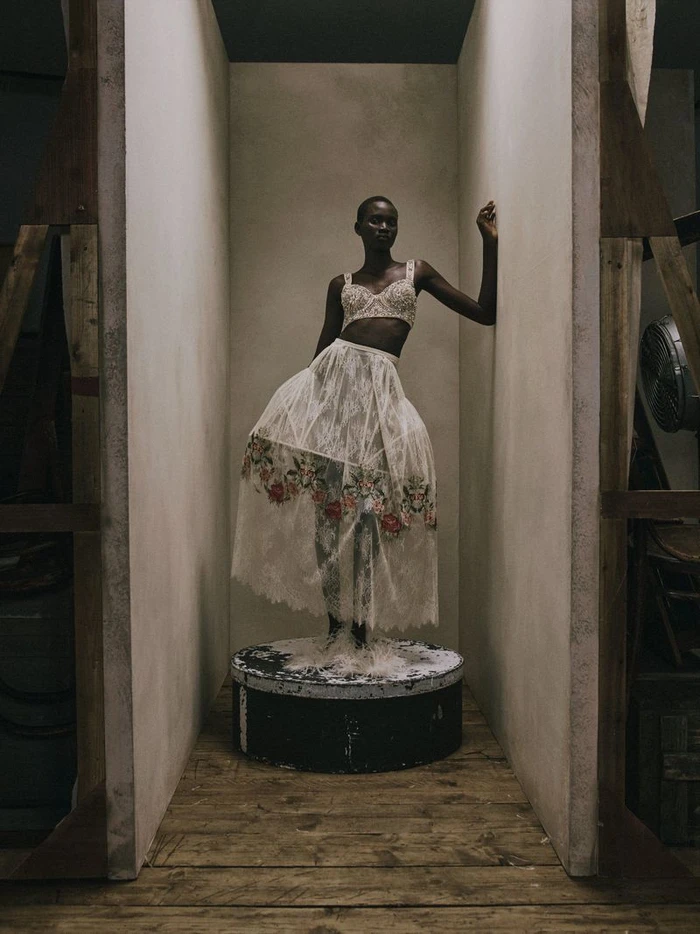 Model posing in a white lace bralette and floral embroidered sheer skirt on a round pedestal inside a studio set.