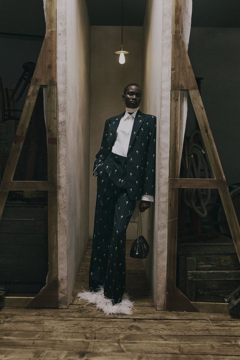 Model Aluel Keror in an embroidered dark tailored suit with white feather-trimmed hems posing on a wooden stage set for Erdem.