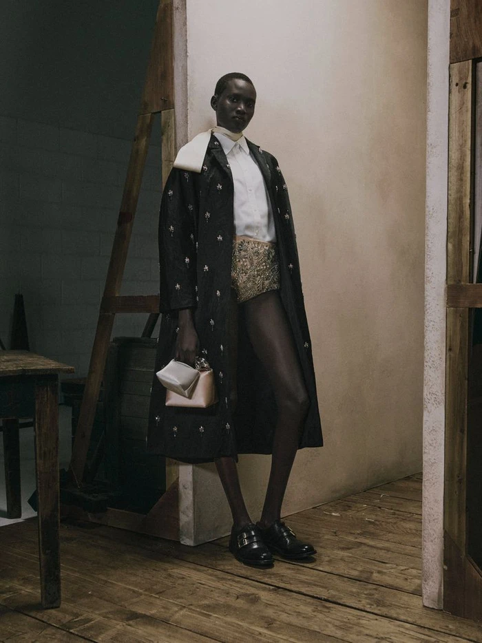 Model Aluel Keror wears an embroidered black coat, white shirt, and brocade shorts standing in an open wooden set doorway.
