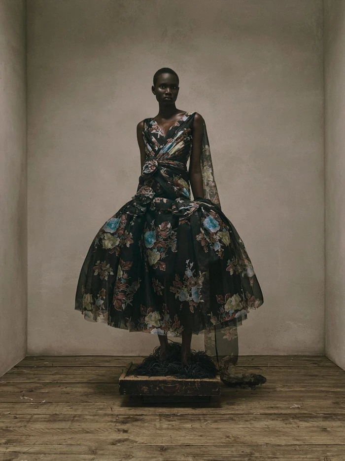 Model wearing a voluminous dark floral sleeveless ballgown while standing on a small wooden platform over rustic plank flooring.