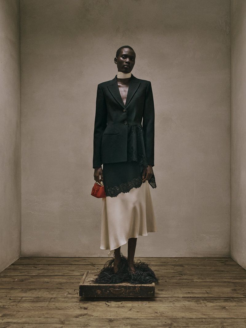Model Aluel Keror wearing a tailored black blazer and cream silk skirt standing on a vintage suitcase in a neutral studio set for Erdem.
