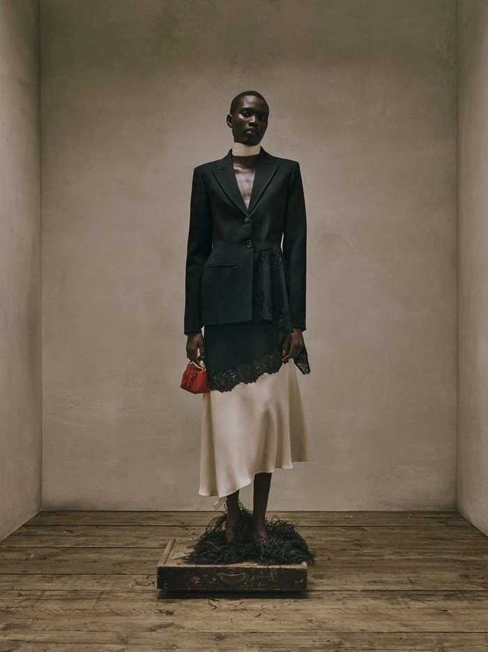 Model Aluel Keror wearing a tailored black blazer and cream silk skirt standing on a vintage suitcase in a neutral studio set for Erdem.