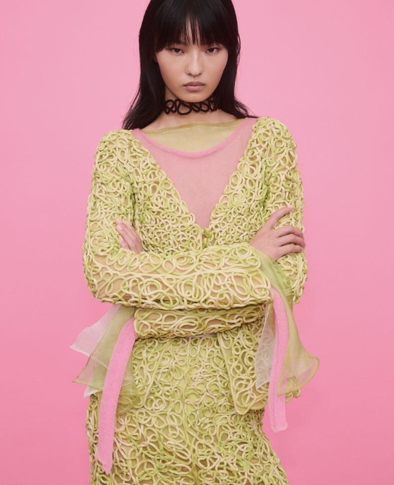 Model with dark hair wearing a textured pale green top with pink trim against a solid pink backdrop