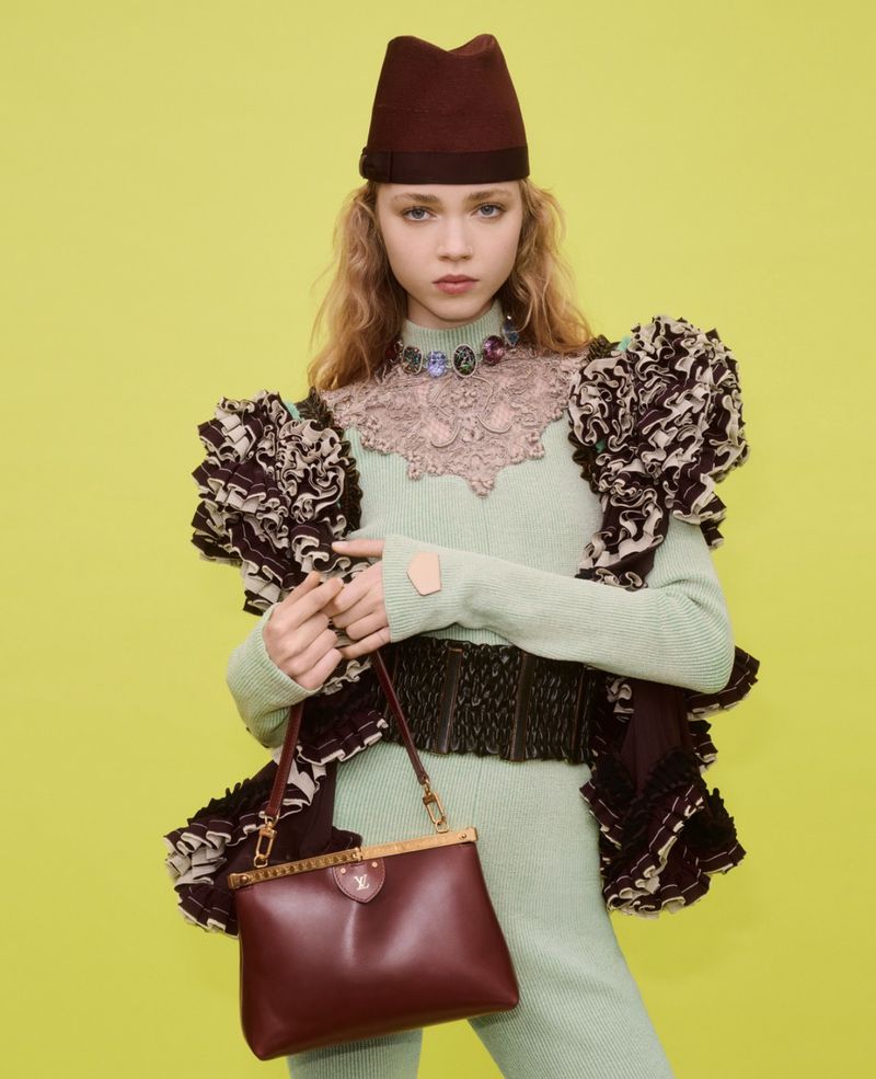 Model wearing a structured burgundy cap, ruffled jacket, and green knit outfit holding a handbag against a yellow background.