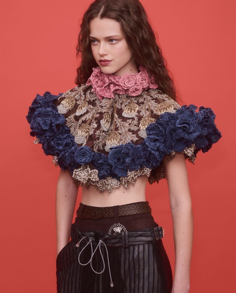 Model wearing a decorative ruffled collar top and patterned cape against a bright red studio background.