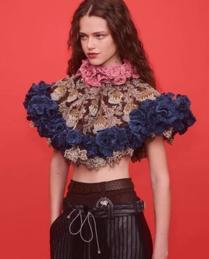 Model wearing a decorative ruffled collar top and patterned cape against a bright red studio background.