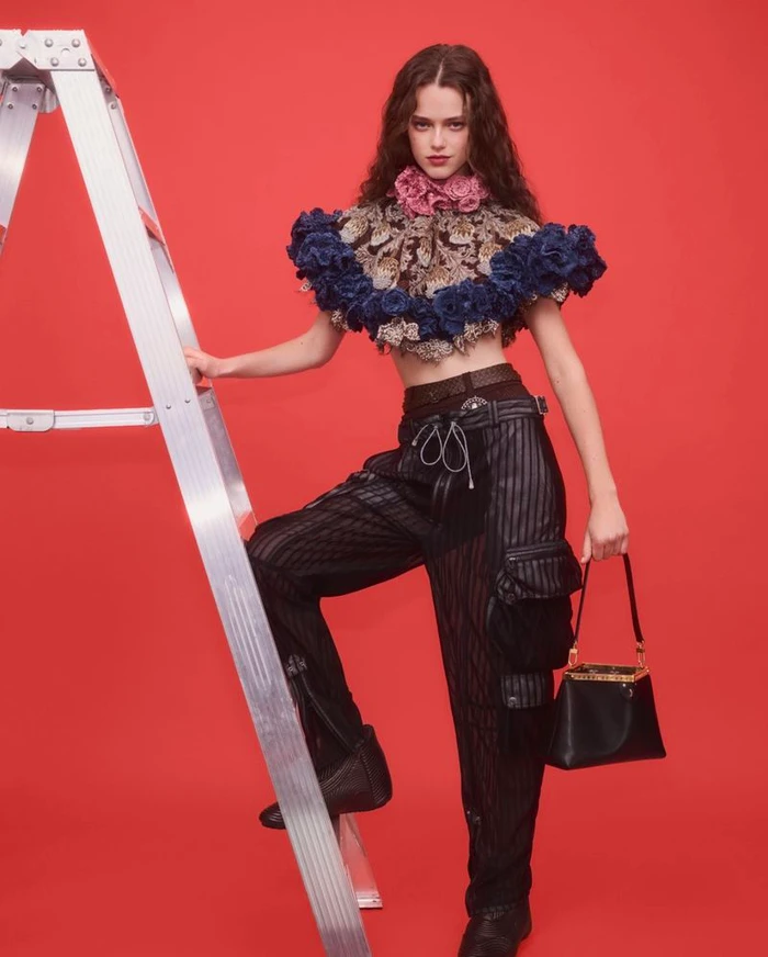 Model poses on a silver ladder in a floral capelet, black cargo pants, and holding a handbag against a red background.