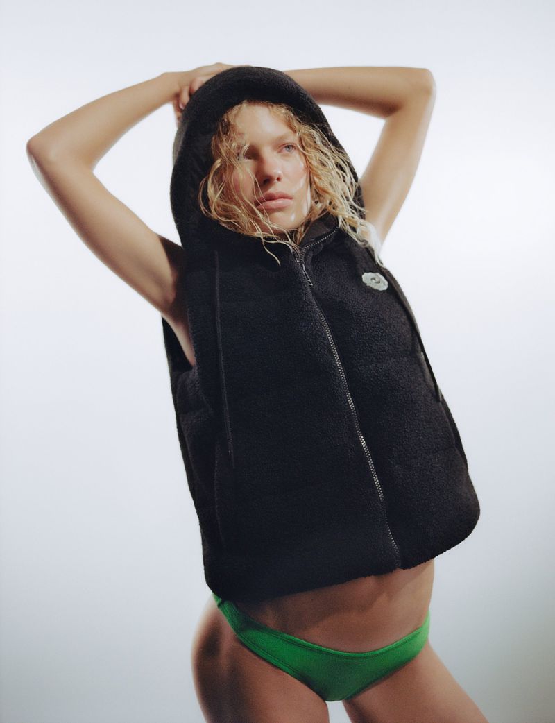 Model posing with hands behind head wearing a black hooded vest and green bottoms against a white studio background.