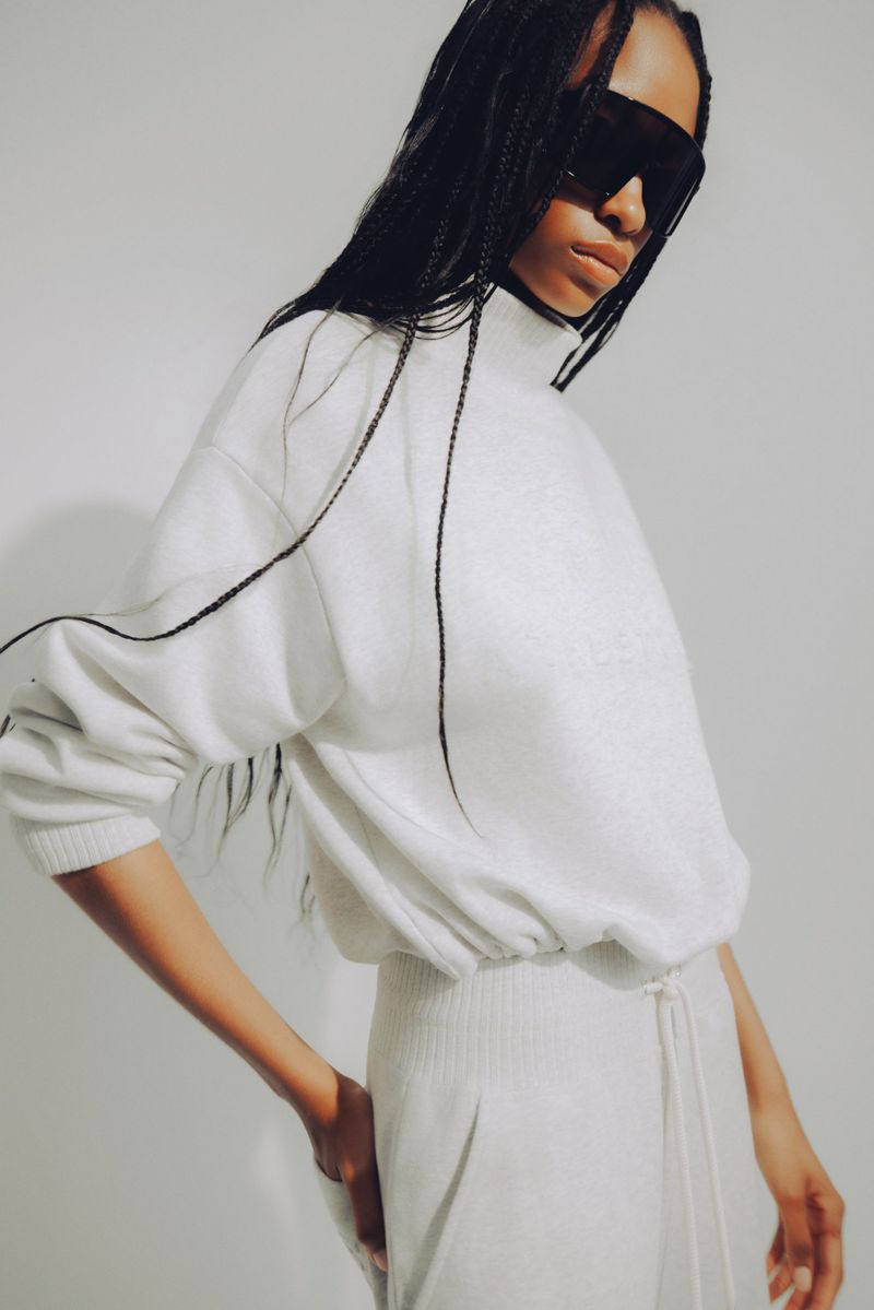 Model with long braided hair wearing oversized dark visor sunglasses and an off-white fleece tracksuit against a plain backdrop.
