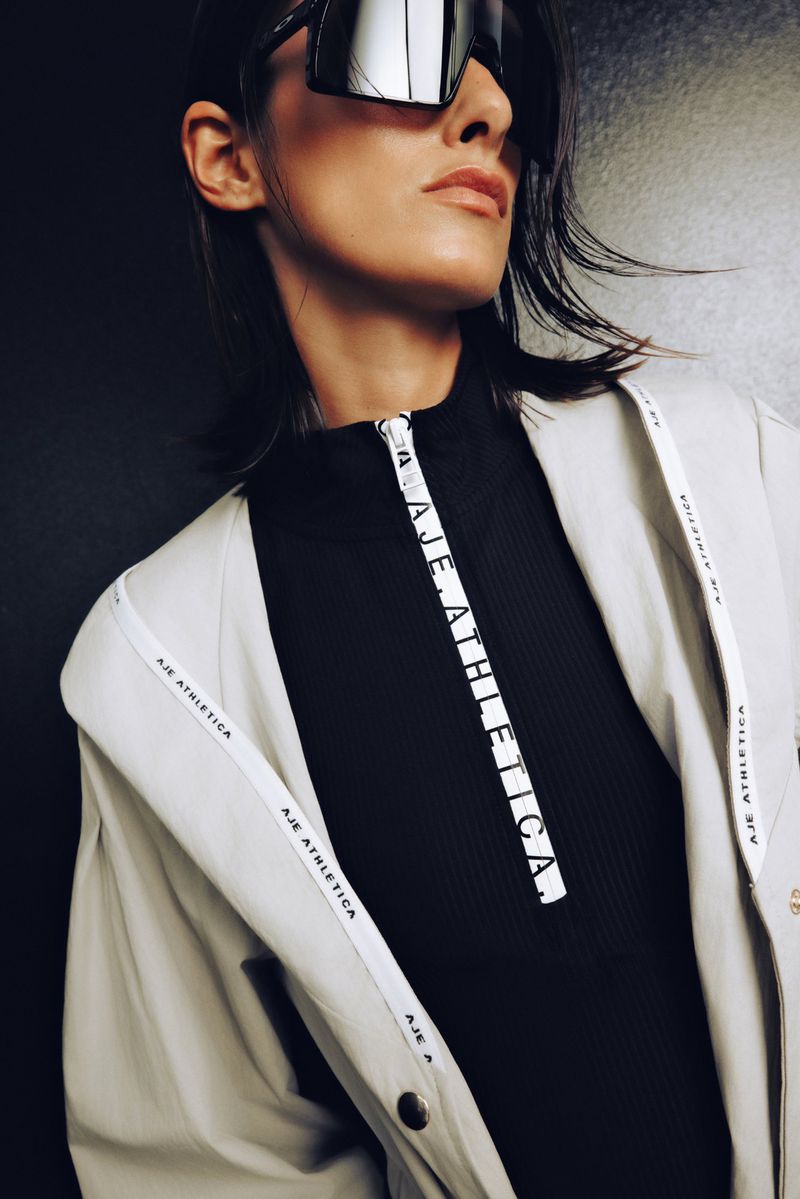 Model wearing oversized sunglasses, a black zipped top, and a light-coloured jacket for the Athletica project by Aje.