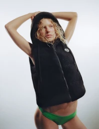 Model posing with hands behind head wearing a black hooded vest and green bottoms against a white studio background.