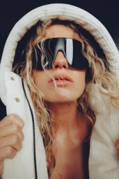 Close-up portrait of a woman wearing oversized black sunglasses and a white hooded jacket with wavy blonde hair framing her face.