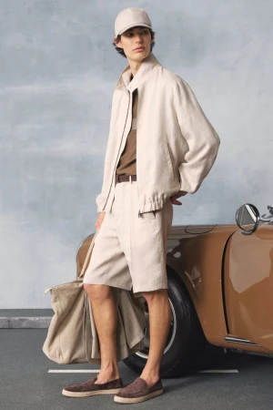 Male model poses in neutral shorts and jacket next to vintage car with a beige tote bag, showcasing a light fashion aesthetic.