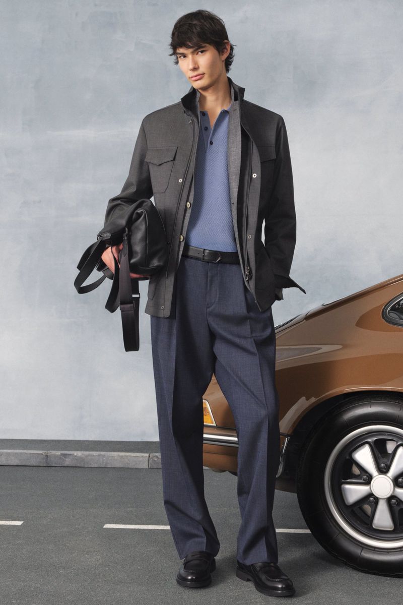 A stylish man poses in a grey jacket, blue polo, and navy pants next to a tan sports car, conveying a blend of classic and modern fashion.