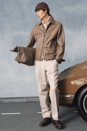 A stylish man poses outdoors wearing a brown leather jacket, beige trousers, and a matching beanie, with a classic car visible.