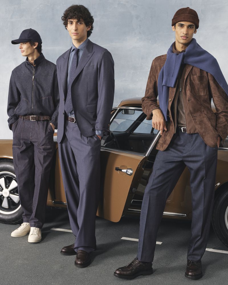 Three male models pose in formalwear, standing in front of a brown vintage car on a paved road under a blue sky. Fashion shoot.