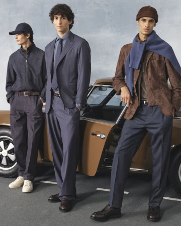 Three male models pose in formalwear, standing in front of a brown vintage car on a paved road under a blue sky. Fashion shoot.