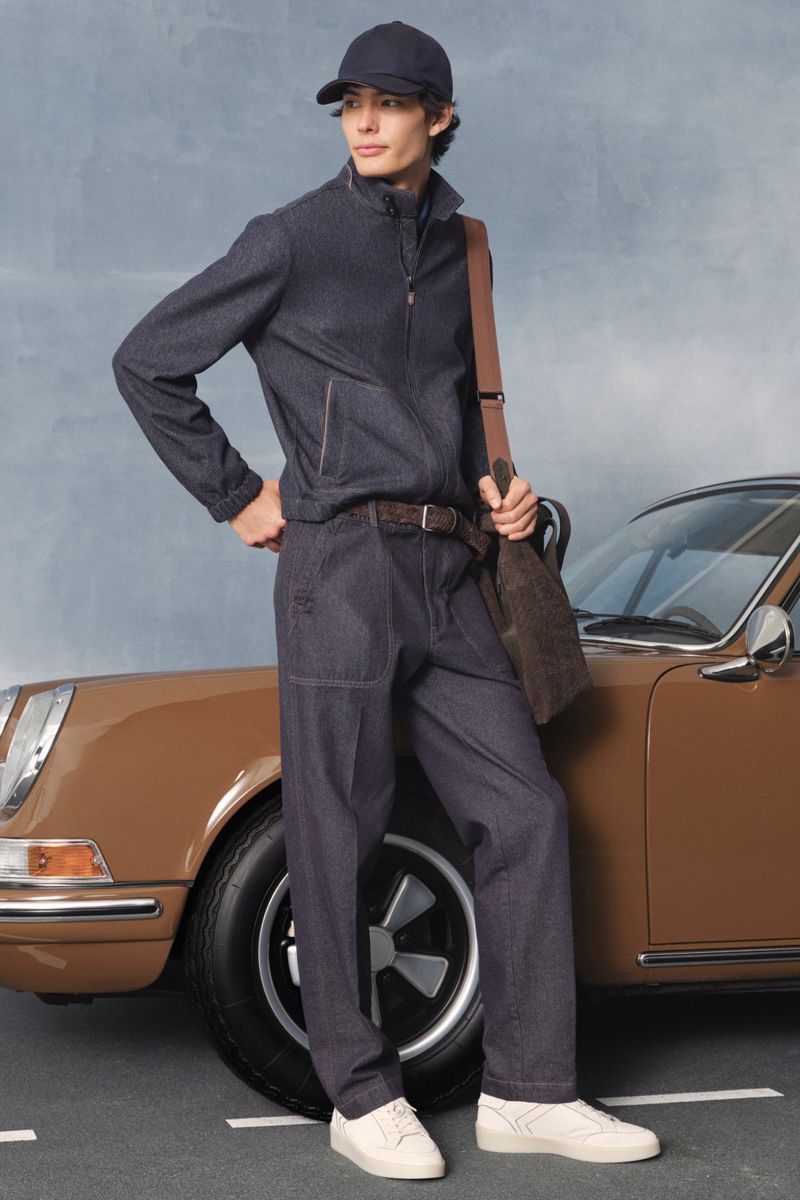Stylish man in navy tracksuit and cap, with shoulder bag and sneakers, poses near a classic brown Porsche on an asphalt road.