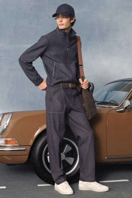 Stylish man in navy tracksuit and cap, with shoulder bag and sneakers, poses near a classic brown Porsche on an asphalt road.
