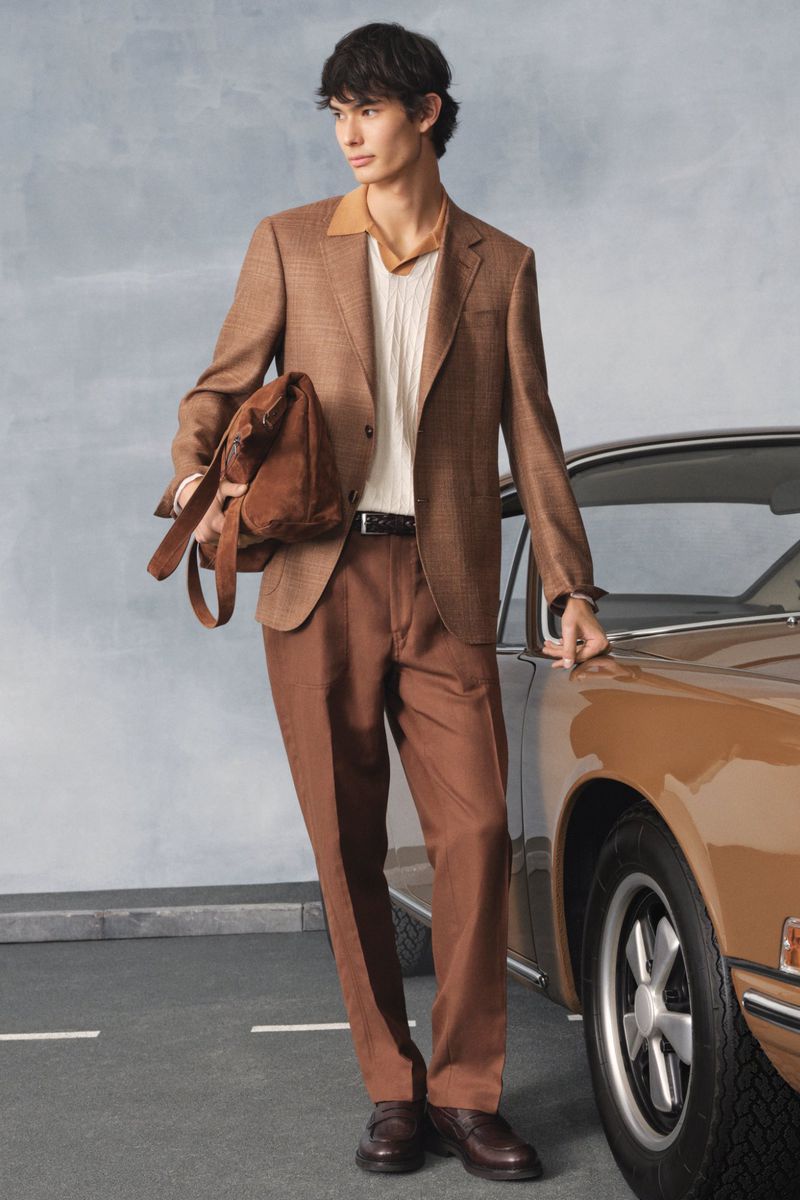 A stylish man in a brown suit stands beside a vintage tan car, showcasing a blend of fashion and automotive elegance in an outdoor setting.