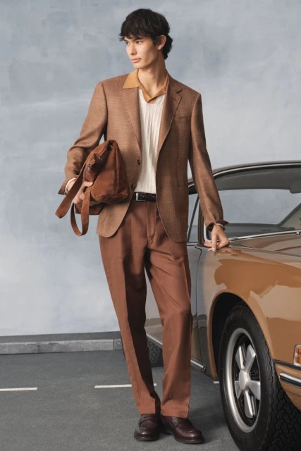 A stylish man in a brown suit stands beside a vintage tan car, showcasing a blend of fashion and automotive elegance in an outdoor setting.