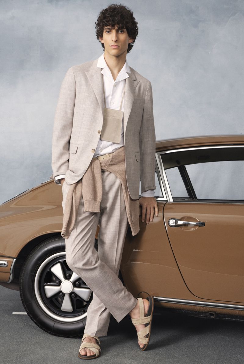 A fashionable male model stands next to a classic brown sports car, wearing a beige suit, white shirt, and tan sandals against a neutral backdrop.