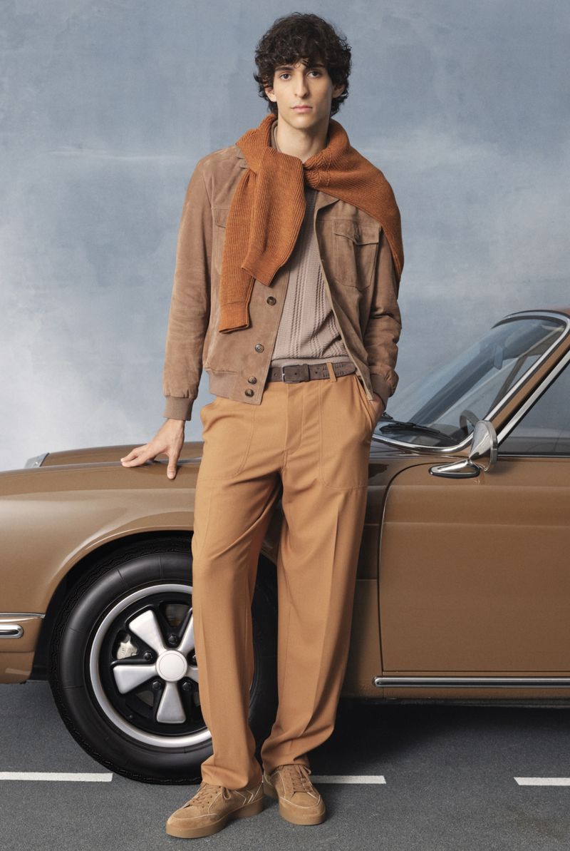 A young adult man posing next to a brown car on an asphalt road, wearing a tan jacket, pants, and shoes with a brown sweater on his shoulders.