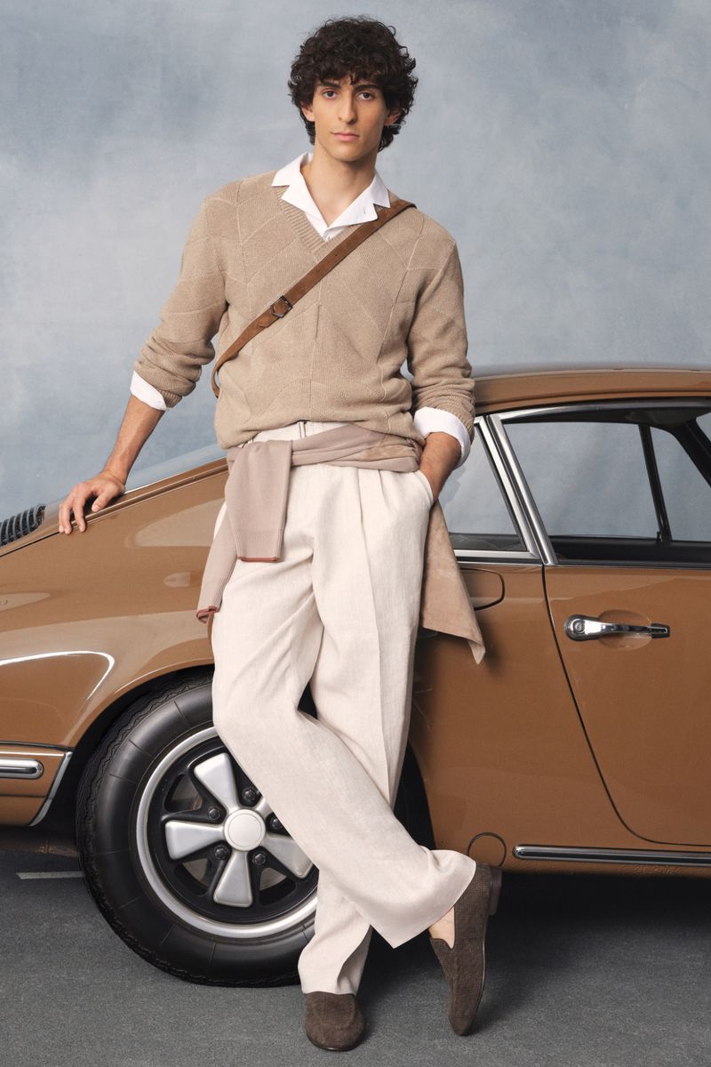 Adult male in a sweater poses next to a vintage brown car. He is wearing neutral-toned pants and leaning against the vehicle.