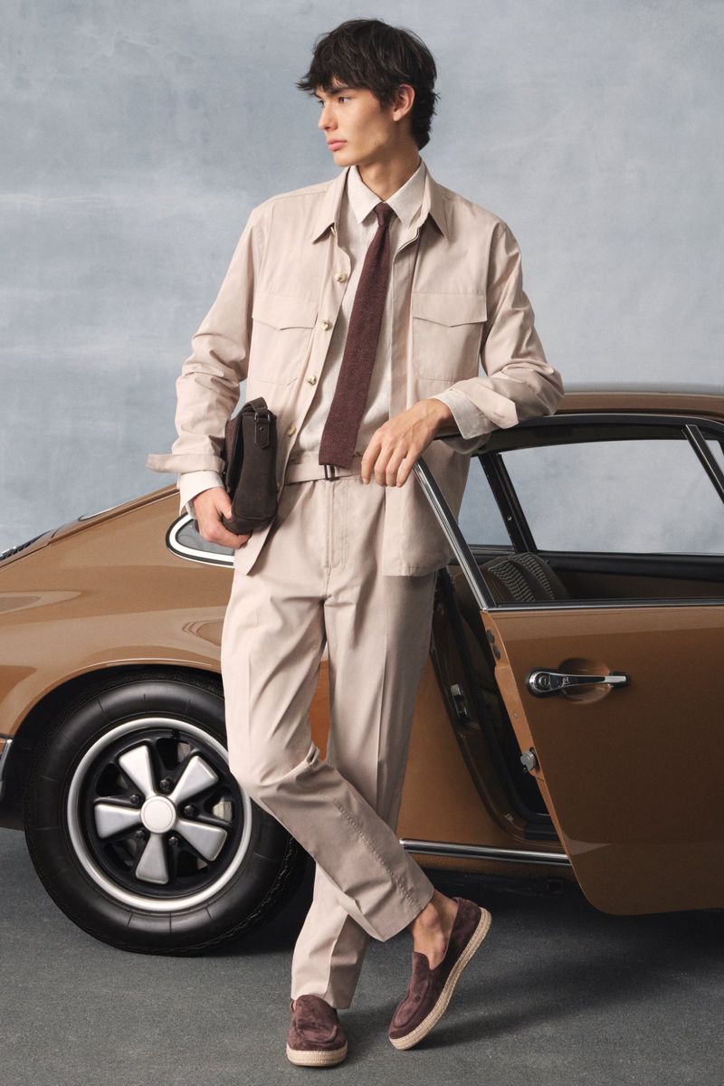 A slender man in beige suit leans against a vintage brown car, looking away from the camera in a studio setting.