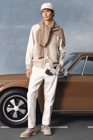 Man in a cream-colored outfit with a patterned scarf poses next to a brown classic car against a light blue backdrop. Fashion editorial.