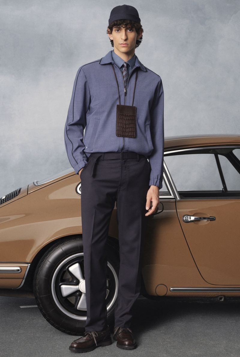 A man in a blue jacket, dark pants, and a cap poses next to a vintage tan car in a studio, showcasing a blend of casual and classic style.