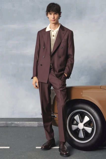 A full-length shot showcases a young adult male model in a plum-colored double-breasted suit and brown loafers, next to a tan-colored car.