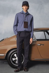 Male model wearing a blue zip overshirt and dark trousers standing next to a vintage brown sports car against a grey backdrop.