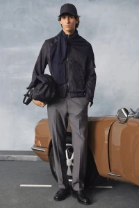 Male model in a dark cap, navy jacket, draped scarf, and grey trousers holding a duffle bag beside a vintage brown car.