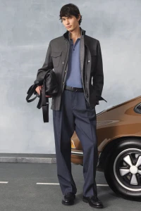Model standing next to a brown vintage car wearing a dark jacket, blue polo, and trousers holding a black bag