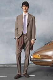 Model standing in a taupe suit and blue tie next to a vintage brown car against a painted studio backdrop