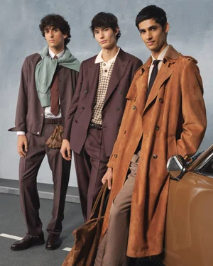 Three male models posing together in tailored menswear looks against a textured grey studio backdrop with a vintage car prop.