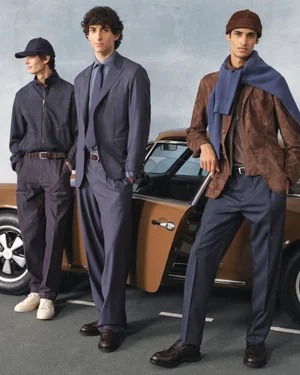 Three male models in tailored menswear and casual jackets posing beside a vintage brown coupe against a textured studio backdrop.