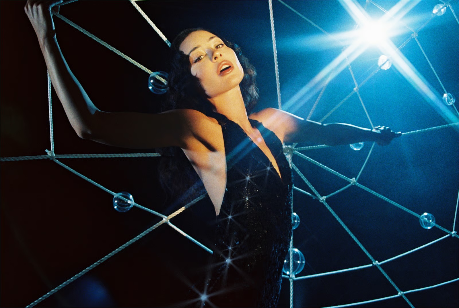Alexa Demie poses in a black plunge dress entangled in a white rope web with glass spheres under dramatic blue backlighting.