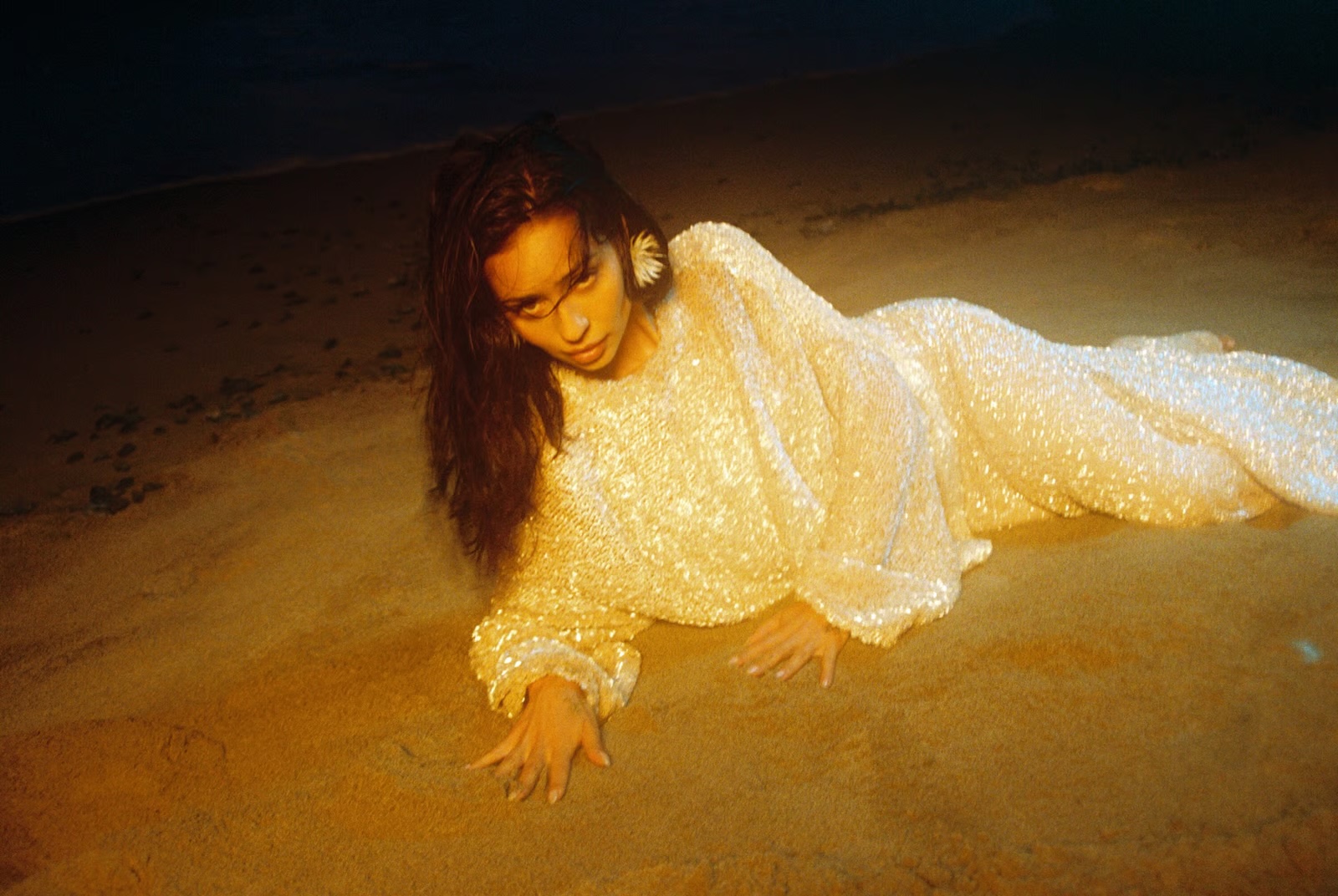 Alexa Demie lying on a sandy beach wearing a shimmering long-sleeved sequined white dress at night.
