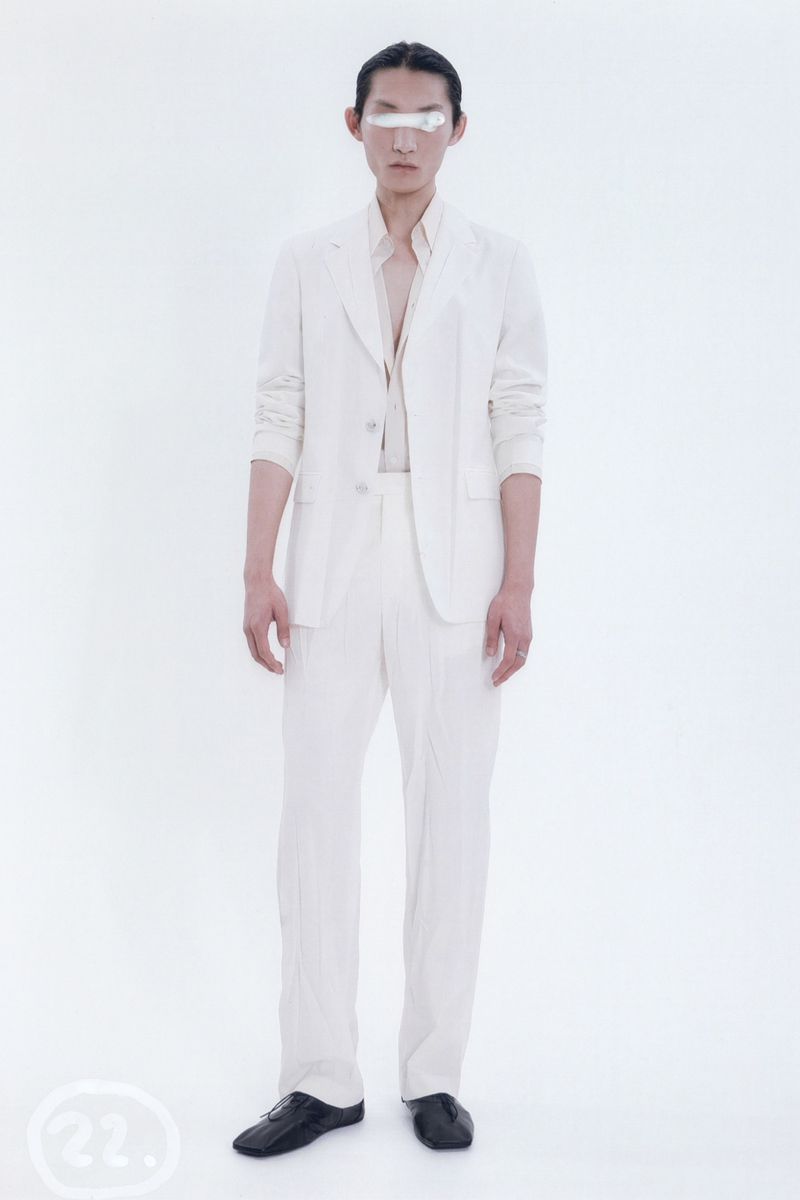 An adult Asian man in a white suit stands in front of a white backdrop wearing black shoes, shot with studio lighting. Minimalist fashion.